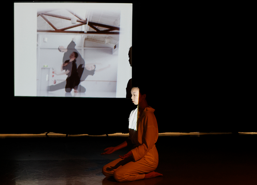 A dancer wearing all-white is kneeled on the floor, arms outstretched, perhaps offering or receiving. Their silhouette against a backdrop projection of faded images of a person dressed in black dancing in a room.