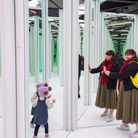 A young child looks at themselves in a mirror maze, standing next to an adult