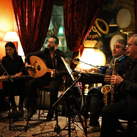 Four musicians from Tarab Ensemble are seated playing music in an intimate setting with lamps and curtains.