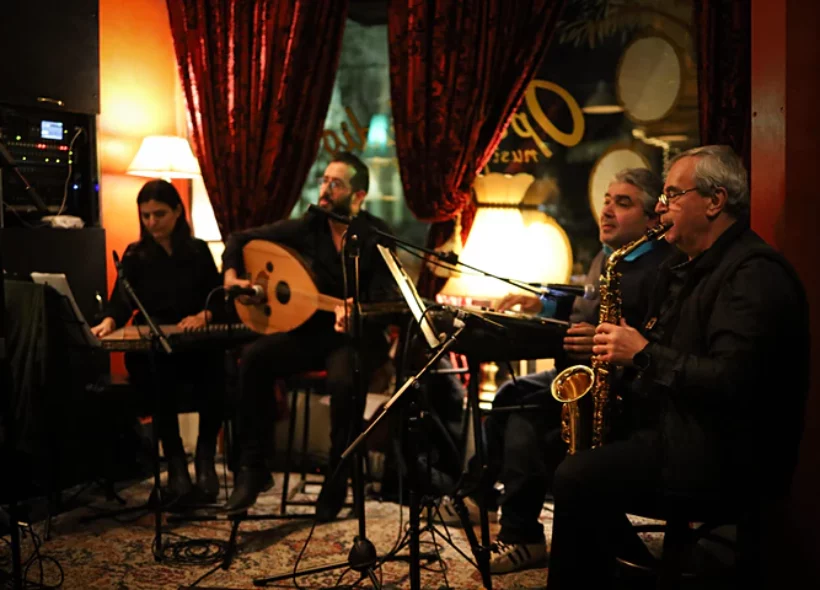 Four musicians from Tarab Ensemble are seated playing music in an intimate setting with lamps and curtains.