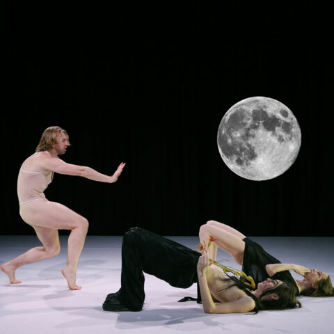 A photo of three performers on a white floor against a black background, featuring an image of the moon. One performer, dressed in a nude tank top with bare legs, stands with arm outstretched, knees bent and back curved, facing the other two performers, one wearing black pants and boots, the other wearing a black short shift dress, who lie on their backs with knees bent.