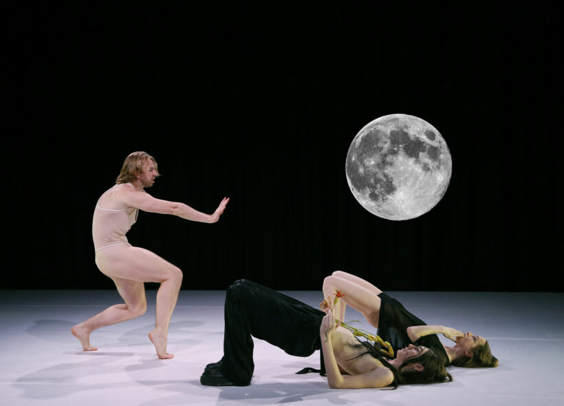 A photo of three performers on a white floor against a black background, featuring an image of the moon. One performer, dressed in a nude tank top with bare legs, stands with arm outstretched, knees bent and back curved, facing the other two performers, one wearing black pants and boots, the other wearing a black short shift dress, who lie on their backs with knees bent.