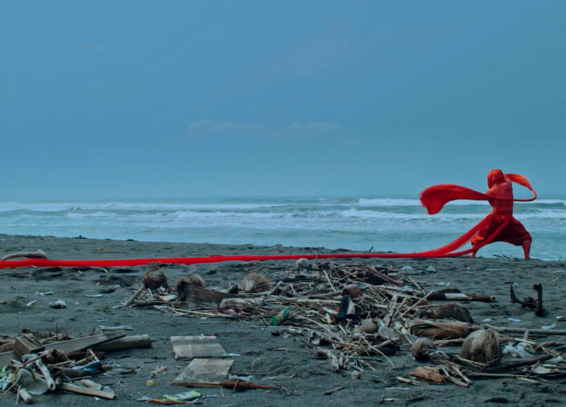 A figure dressed and painted in red is dancing on the foreshore of Parang Kusumo beach in Jogjakarta, Indonesia, revered as a sacred place in Javanese culture. They are twirling a long sash of red fabric, which is wrapped around their head and waist and trails long behind them on the sand.