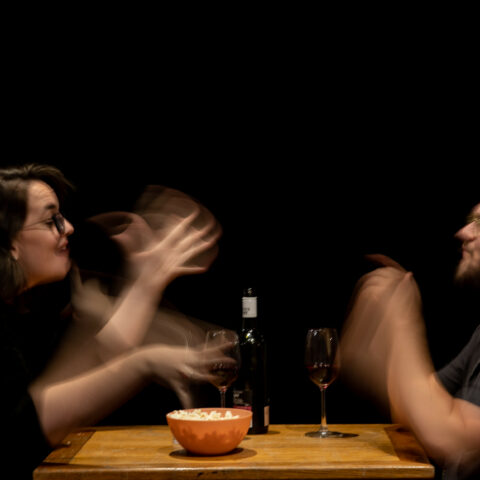 Two people sit at a small dining room table intimately signing to one another in Auslan over popcorn and red wine. Their gestures are captured in slow motion, blurring in speed and emotion as they communicate.  