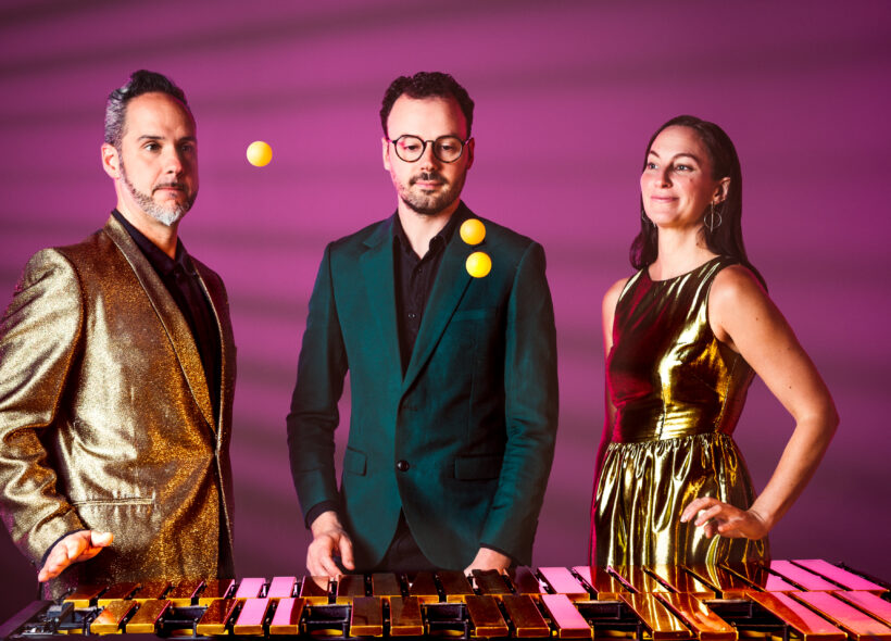 Both the background and the golden bars of the marimba are radiated by a beautiful pinkish magenta. Three musicians stand behind an instrument: on the left, a white man with a silver wave of hair and finely shaped goatee; in the middle a white man with dark brown hair, short cut beard and black circular rimmed glasses; and on the left a white long brown haired woman with dangling circular earrings. All three stare transfixed at three fluorescent ping pong balls either descending or ascending, above the instrument below. They each wear clothes for a party or a talent contest: the man on the left sports a confetti golden jacket; the woman on the right a shiny golden sleeveless dress. Together they flank the man wearing a jade regal green formal jacket.