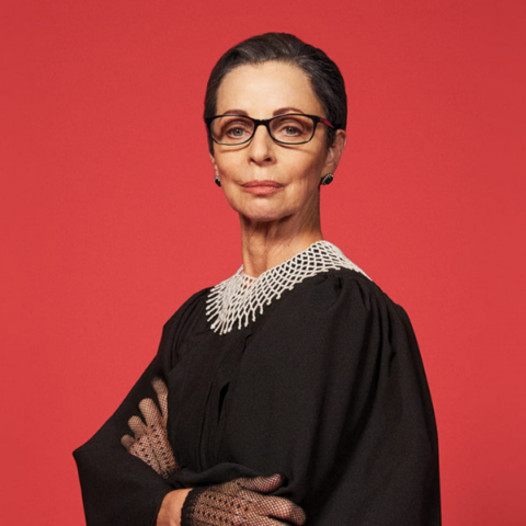 Woman in glasses wearing a judge's gown and gloves standing sternly upon a red background.