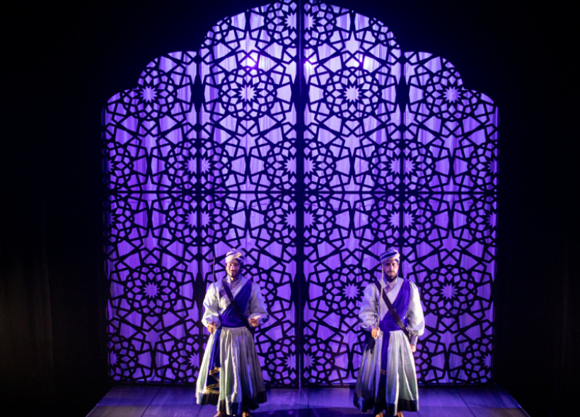 Two guards in purple and white garments in front of a large stain glassed purple glass