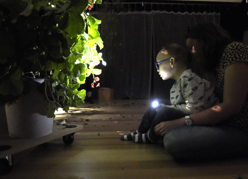 A Sensory Play photo. A child sits on their adult's lap on a wooden floor in a darkened space. They are pointing a small torch/flashlight at a large potted plant on a low trolley. Photographer: Carla Gottgens, Polyglot Theatre