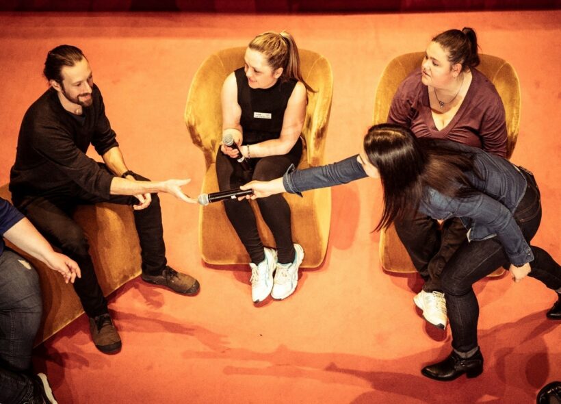 Birds Eye view of 5 people ranged on theatre armchairs. A microphone is being handed to one person from another.