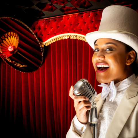 Performer in a white top hat and white tuxedo posing in front of theatre curtains with a vintage microphone in hand.