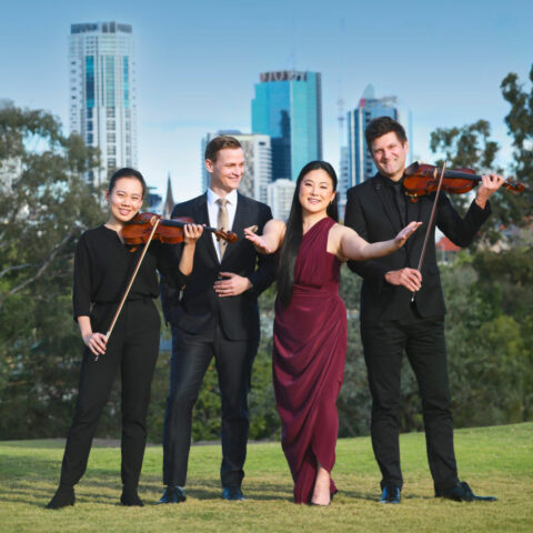 Four people stand on a grassy landscape with skyscrapers in the background. The people on the far left and far right hold violins. The lady second from the right holds her arms outstretched to the camera whilst the man second from the left looks at her. 