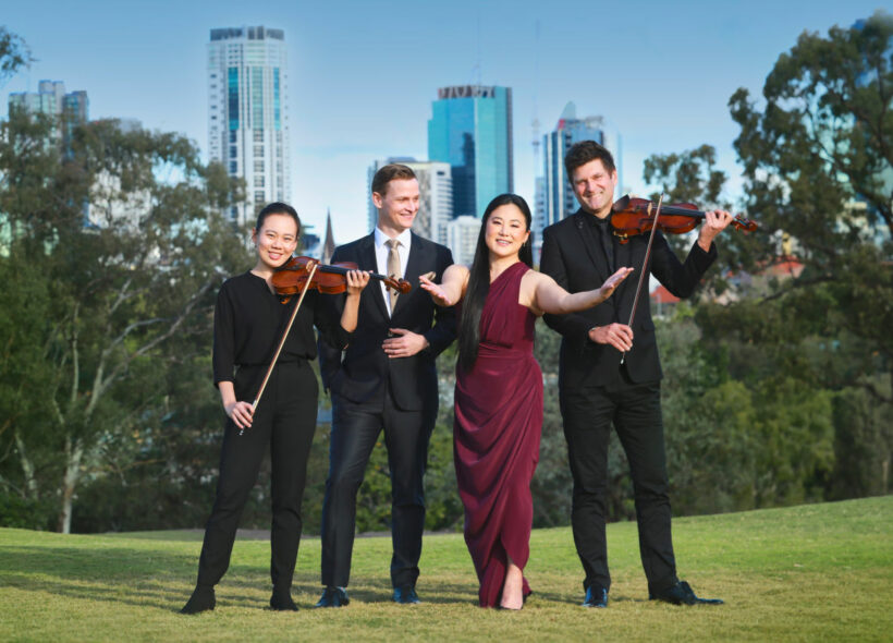 Four people stand on a grassy landscape with skyscrapers in the background. The people on the far left and far right hold violins. The lady second from the right holds her arms outstretched to the camera whilst the man second from the left looks at her. 