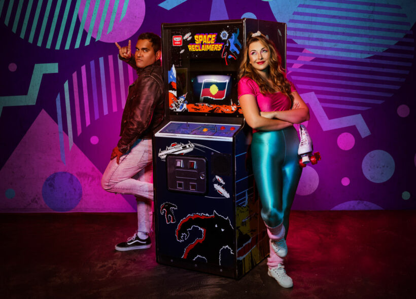 Two people stand in front of a patterned background. The person on the left leans against an arcade game looking at the camera, while the person on the right leans against the arcade game looking up, holding one roller skate in her hand. The arcade game is space reclaimers. 