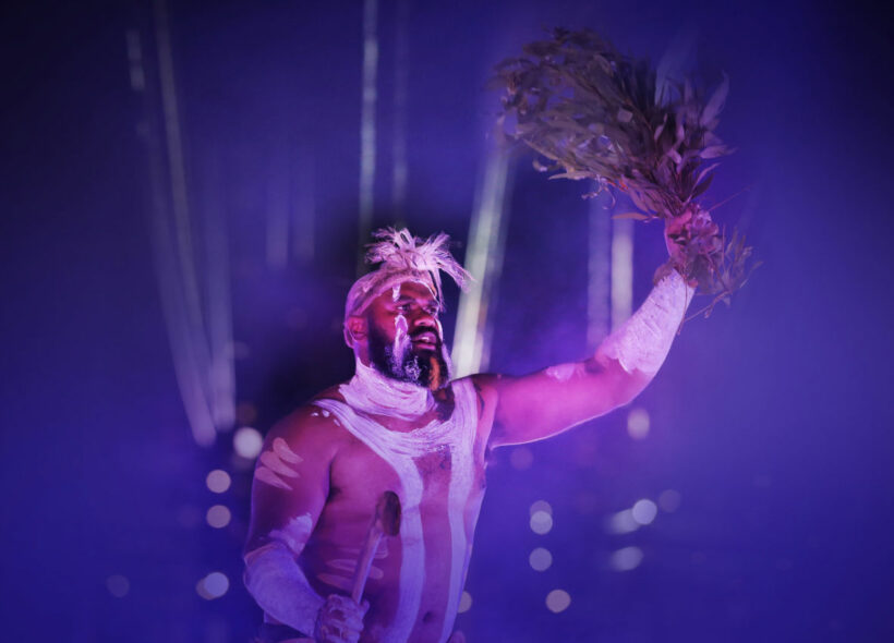 An Indigenous man is centred, covered in traditional attire including white body paint. He holds branches with leaves in his hand in the air. 