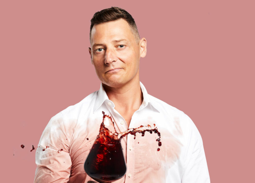 A man stands in front of a pink background. He is wearing a white shirt that is partially soaked with red wine. In front of him, he holds a wine glass that seems to be swirling, with red wine splashing over the edges.  
