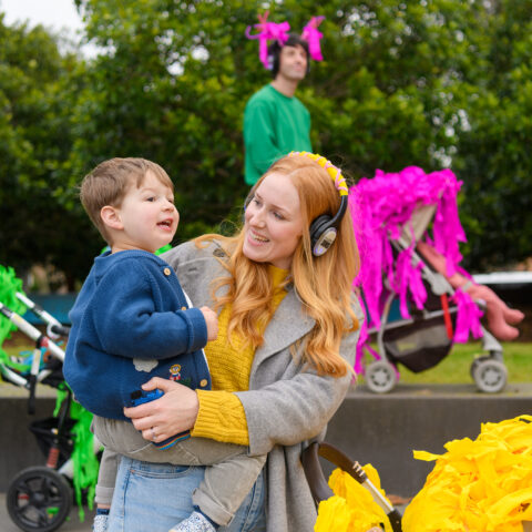 A Pram People photo. An adult in a yellow jumper and headphones holds their child, smiling at them. Other adults with small children and brightly decorated prams stand in the background. They are outside on grey steps, with green trees in the background. Photographer: Sarah Walker