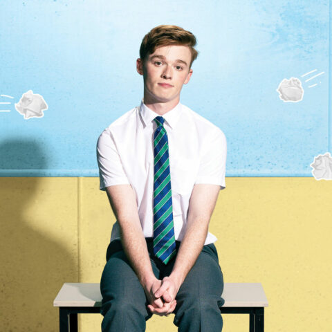 A young boy sits on a desk in front of a blue and white wall. He is dressed in a white button up, black pants and a blue and green striped tie. Five animated scrunched up paper balls seem to be thrown at him. 