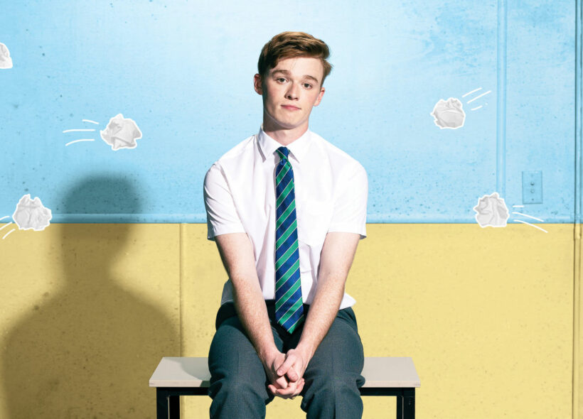 A young boy sits on a desk in front of a blue and white wall. He is dressed in a white button up, black pants and a blue and green striped tie. Five animated scrunched up paper balls seem to be thrown at him. 