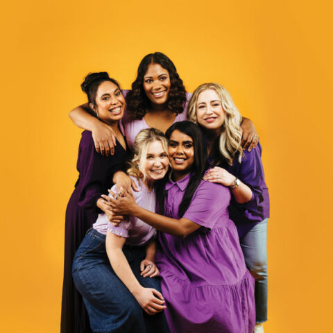 Five women dressed in various shades of purple stand in front of an orange background. They face the camera smiling, holding each other in an embrace. 
