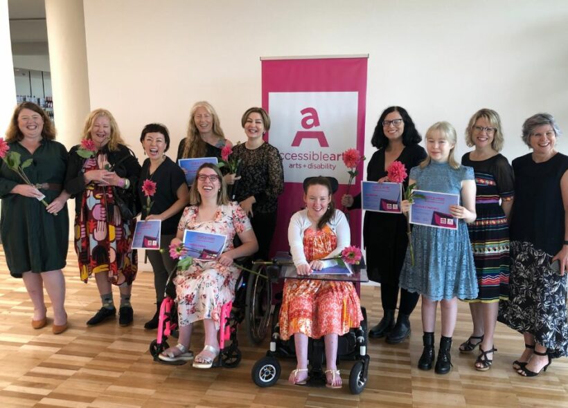  A group of women are in front of a pink Accessible Arts banner. They are smiling and holding pink flowers and certificates.
