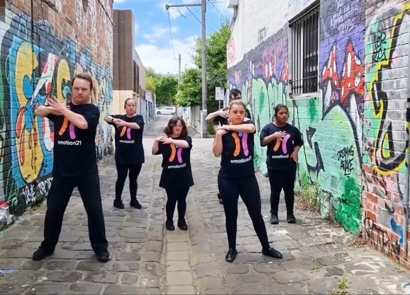 People with down syndrome wearing black uniforms dancing in a Melbournian Alley with colourful street art.