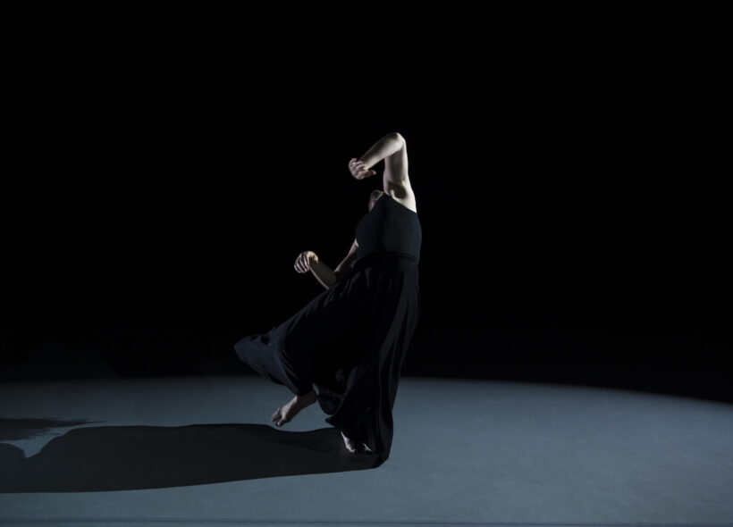 A single dancer dressed in all black leans backward with their face out of view