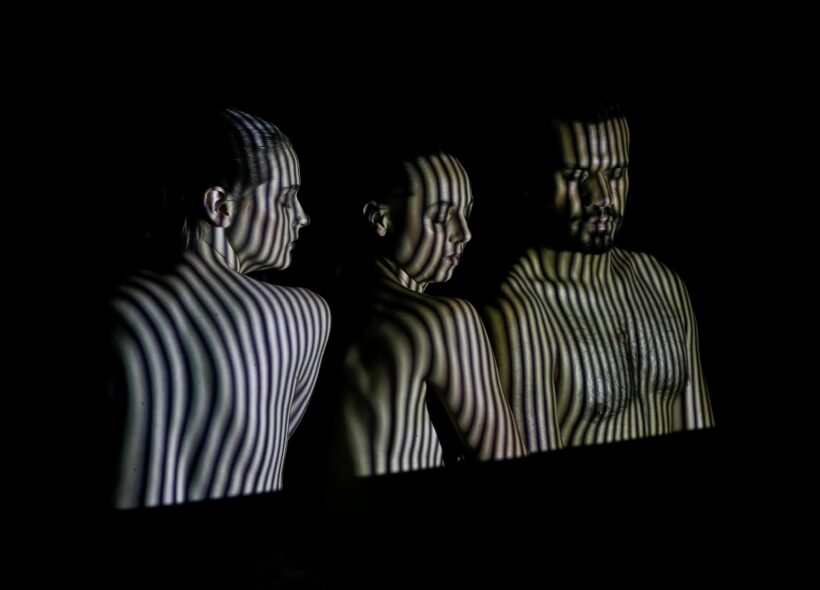 Garabari - Image by Jeff Busby - A photo of three Indigenous dancers side-by-side with their eyes closed, against a black background. Dark vertical stripes are projected across their faces and bare skin. 