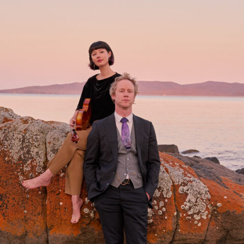 Woman sitting with a viola on a boulder in front of a river mouth and man leaning up against the same boulder