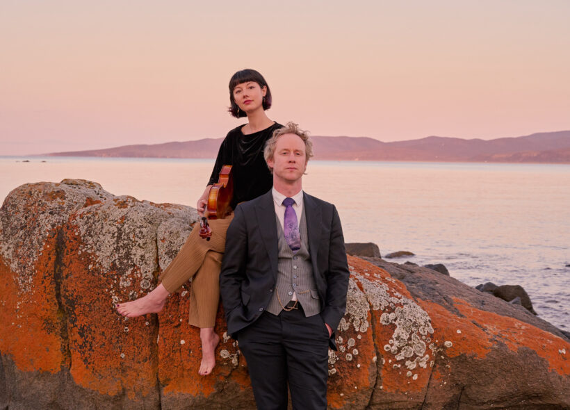 Woman sitting with a viola on a boulder in front of a river mouth and man leaning up against the same boulder