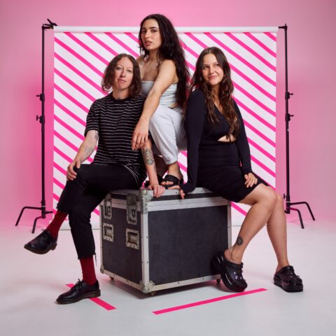The three band members from Camp Cope sit on top of a road case, with a striped pink background.