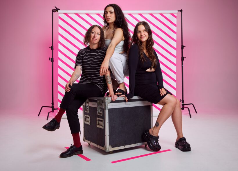 The three band members from Camp Cope sit on top of a road case, with a striped pink background.