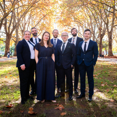 Seven men and women dressed in navy suits and dresses standing in a park and surrounded by large autumn trees