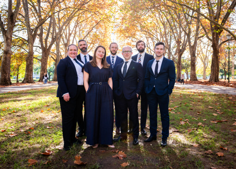 Seven men and women dressed in navy suits and dresses standing in a park and surrounded by large autumn trees