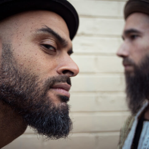 Close up of the side of a man's face, with a beard and hat, and another man in the background facing the opposite way