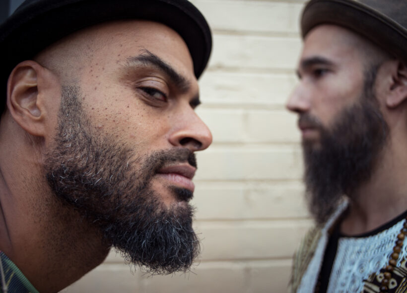 Close up of the side of a man's face, with a beard and hat, and another man in the background facing the opposite way