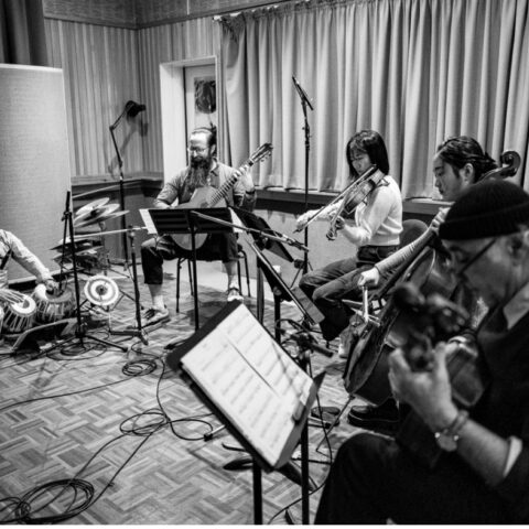 Black and white image of musicians in a recording studio with guitars, violin, cello and tablas