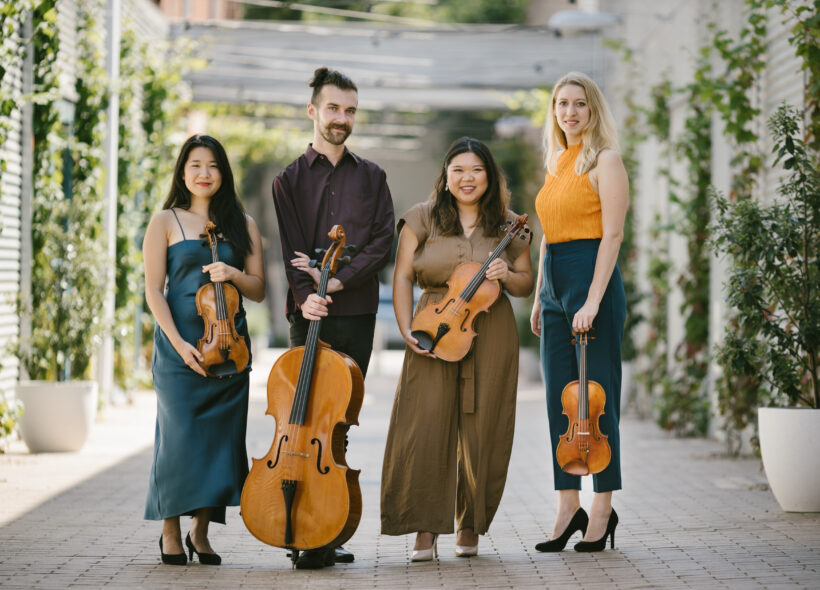 four musicians standing in a walkway surrounded by trees, with their string instruments