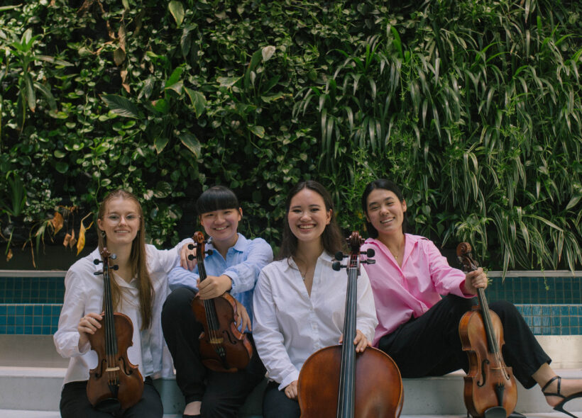Four women in black pants and shirts sitting in front of a pool with string instruments