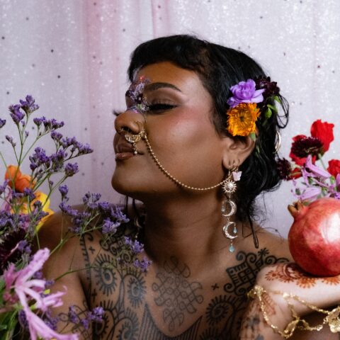A woman wearing traditional Indian jewelry and henna on her skin, holding a pomegranate and surrounded by colourful flowers