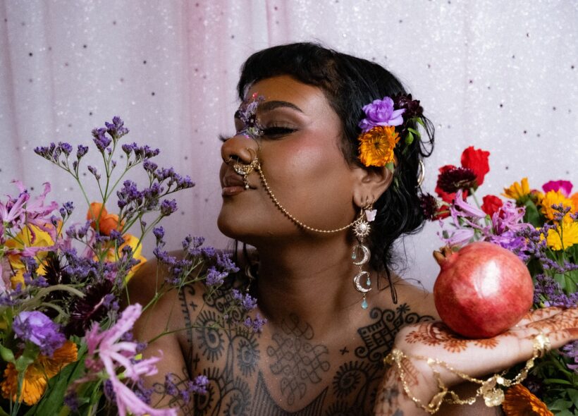 A woman wearing traditional Indian jewelry and henna on her skin, holding a pomegranate and surrounded by colourful flowers