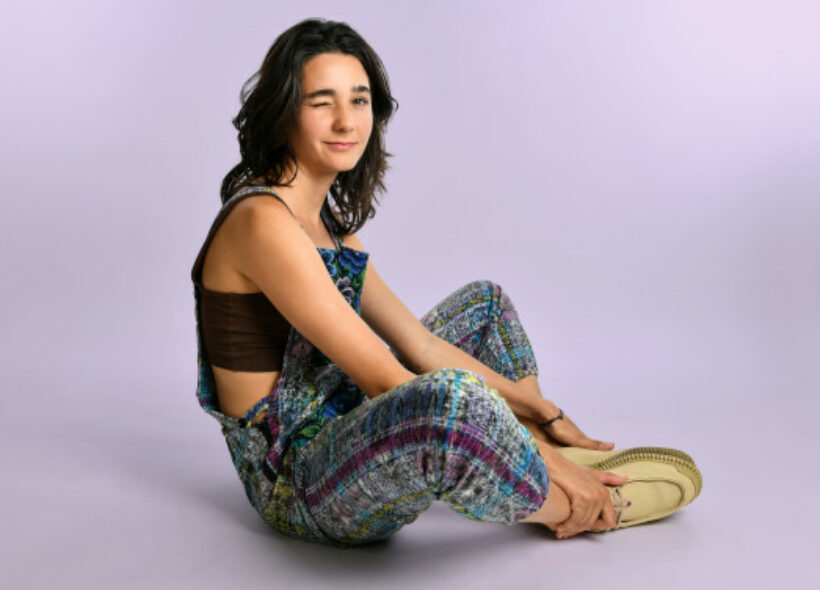 Lara Ricote sitting on the ground in patterned overalls winking at the camera.