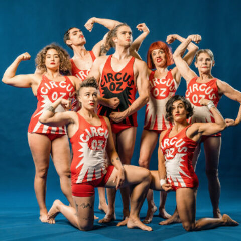 Group of seven performers in red and white Circus Oz outfits flexing.