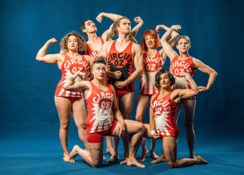 Group of seven performers in red and white Circus Oz outfits flexing.