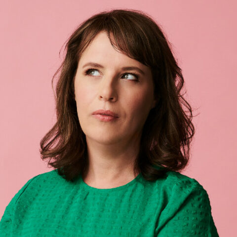 Anne Edmonds shown in a green top in front of a salmon pink background, looking out of the corner of her eye.