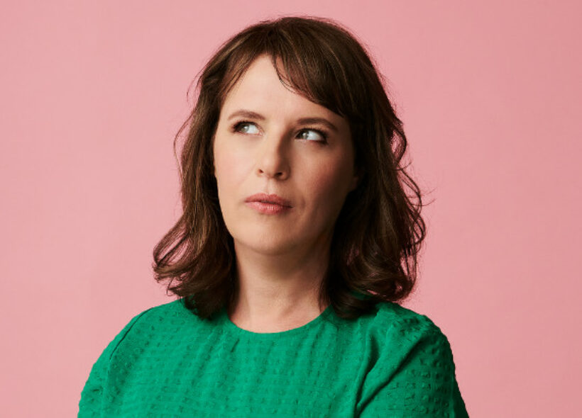 Anne Edmonds shown in a green top in front of a salmon pink background, looking out of the corner of her eye.