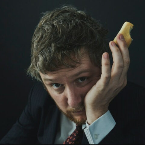 Tim Key in a suit and tie with his cheek in his hand, holding a snack between two fingers.