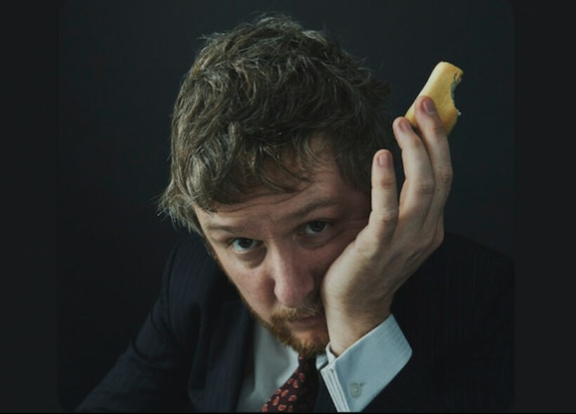 Tim Key in a suit and tie with his cheek in his hand, holding a snack between two fingers.