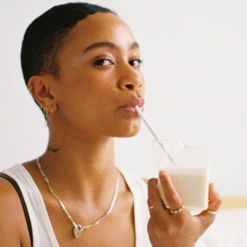 Aurelia St Clair in a white top in front of a white background sipping a glass of milk or non-dairy beverage with a straw.