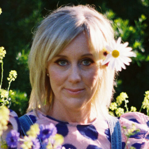 Claire Hooper outdoors surrounded by little green flowers with a daisy behind her ear.