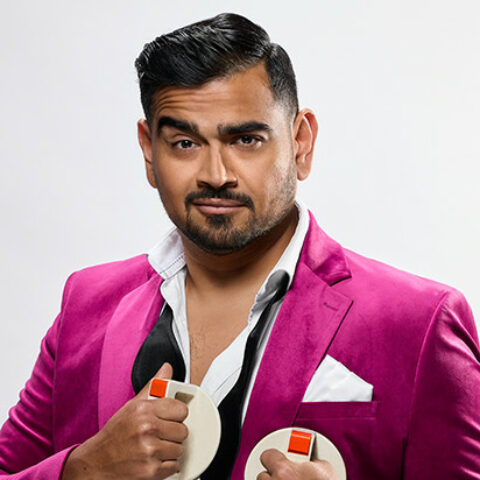 Dilruk Jayasinha in a pink suede suit, an undone tie around his neck, holding defibrillator paddles against his chest. and 